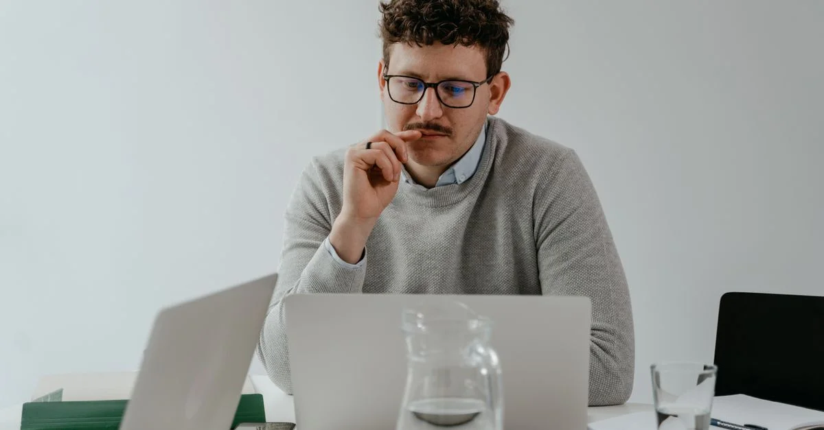 Homme en lunettes travaillant attentivement sur un ordinateur portable à un bureau avec des papiers et une bouteille d'eau sur la table.