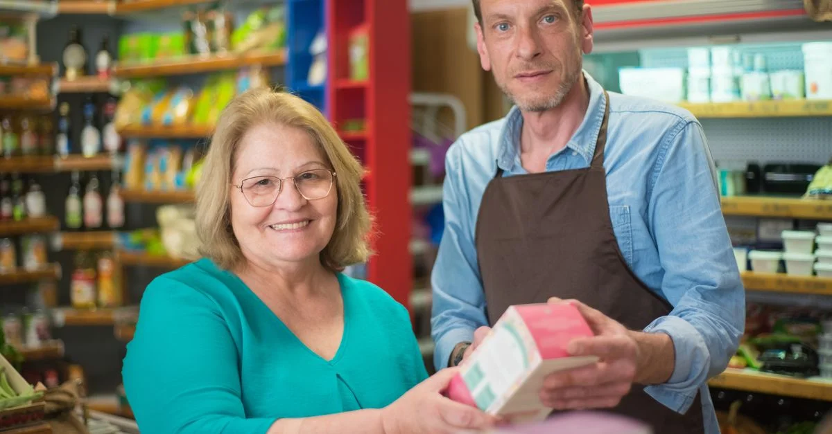 Une femme senior et un commerçant souriant lors d'une interaction dans une épicerie au Portugal.