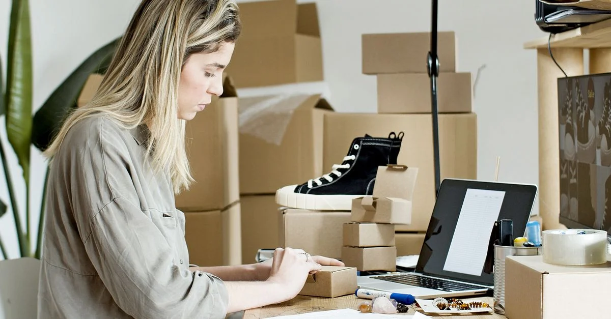 Une femme en train de préparer des boîtes pour sa boutique en ligne, entourée de matériaux d'emballage et d'un ordinateur portable.