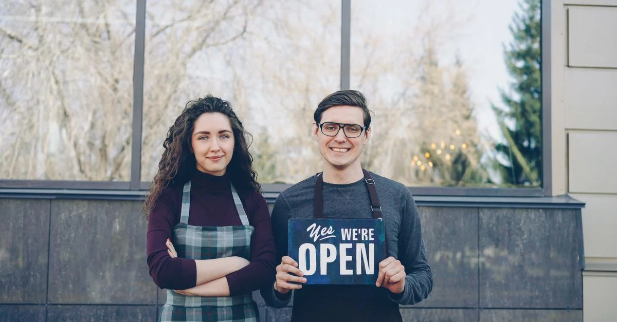 Deux propriétaires de commerce se tenant à l'extérieur, tenant un panneau 'Oui, nous sommes ouverts', accueillant les clients.