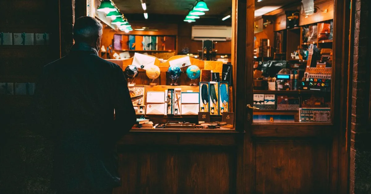 Un homme contemple la vitrine d'une boutique à l'éclairage chaleureux, où sont exposés divers articles colorés.