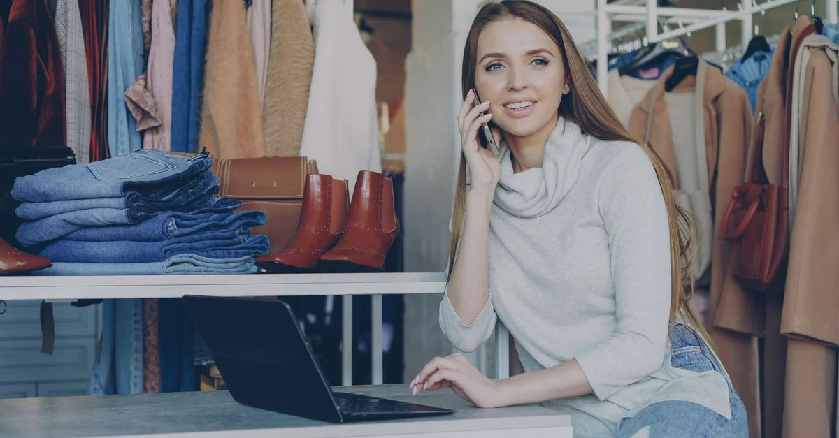 Femme dans une boutique parle au téléphone avec un ordinateur portable, entourée de vêtements.