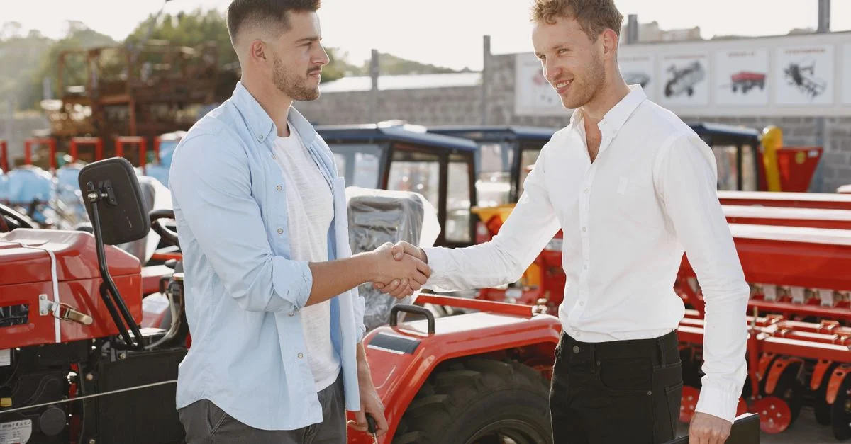 Deux hommes se serrant la main près d'un tracteur, symbolisant un accord commercial ou un contrat d'achat, en extérieur.